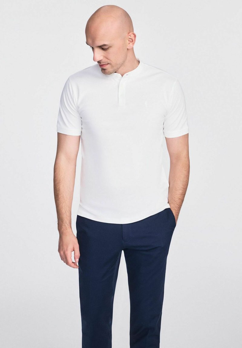 Biała koszulka polo z krótkim rękawem i kołnierzykiem typu henley, w zestawieniu z granatowymi spodniami. Gładka faktura, minimalistyczny design i subtelny detal logo.