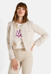 Femme avec des cheveux bruns raides portant une veste beige courte, un pantalon clair et un sweat-shirt blanc avec des lettres violettes.