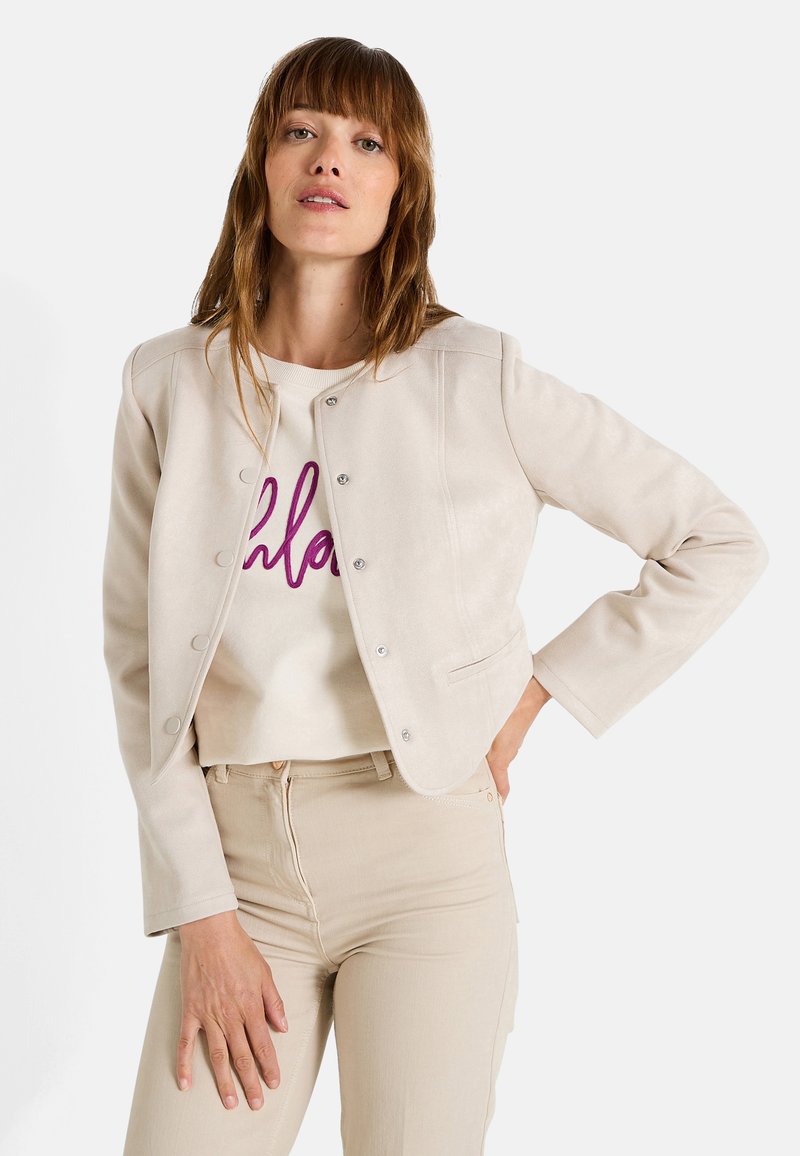 Femme avec des cheveux bruns raides portant une veste beige courte, un pantalon clair et un sweat-shirt blanc avec des lettres violettes.