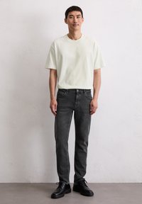 Jonge man die tegen een witte muur staat, gekleed in een wit T-shirt, donkergrijze jeans en zwarte veterschoenen, met een neutrale uitdrukking recht vooruit kijkend.