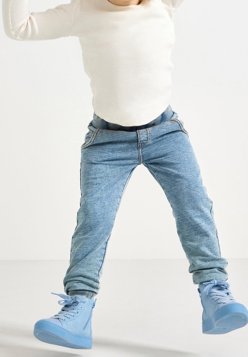 Enfant portant des baskets bleu clair, un jean bleu clair et une chemise à manches longues blanche, sautant les jambes écartées sur un fond blanc uni.