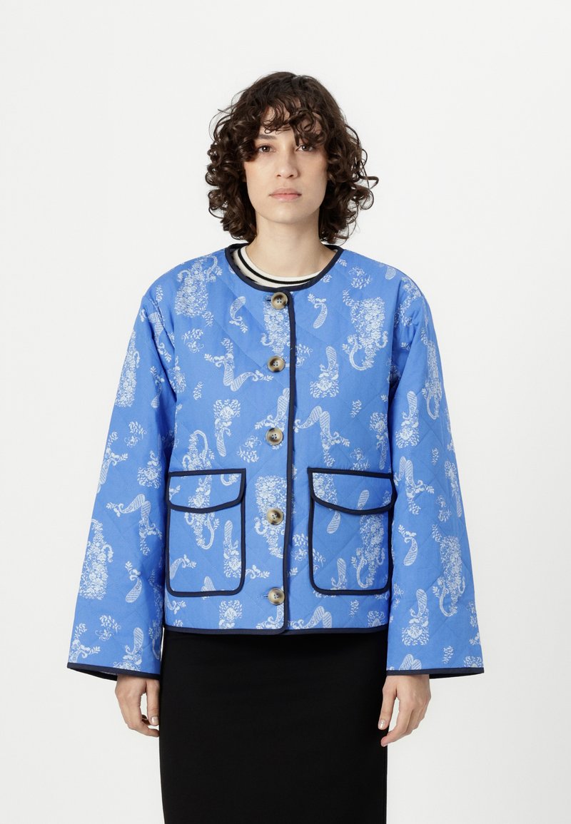 Blue quilted jacket with white floral patterns, featuring two front pockets, round neckline, and black piping along edges and pockets.
