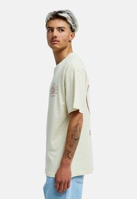 Jeune homme aux cheveux décolorés, portant un t-shirt beige à motif graphique et un jean bleu clair, regardant vers sa gauche, montrant un tatouage détaillé sur l'avant-bras.
