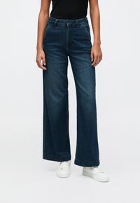 Pessoa vestindo jeans de perna larga de cintura alta, escuros e sapatos brancos, de pé contra um fundo claro e liso.