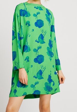 Løs, grøn kjole med lange ærmer med abstrakte mørkeblå blomsterprint, båret af en person med hænderne afslappet ved siden.