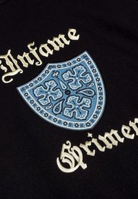 Zwarte stof met een geborduurd blauw schildontwerp met ingewikkelde patronen, verfraaid met gouden letters die "Artfame Grimen" zeggen.