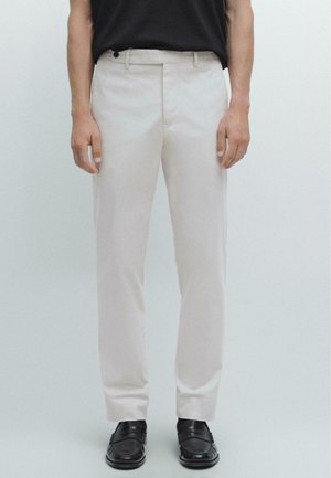 Pantaloni beige chiaro con vestibilità dritta, dotati di chiusura con bottone e tasche laterali. Abbinati a mocassini neri su uno sfondo semplice.