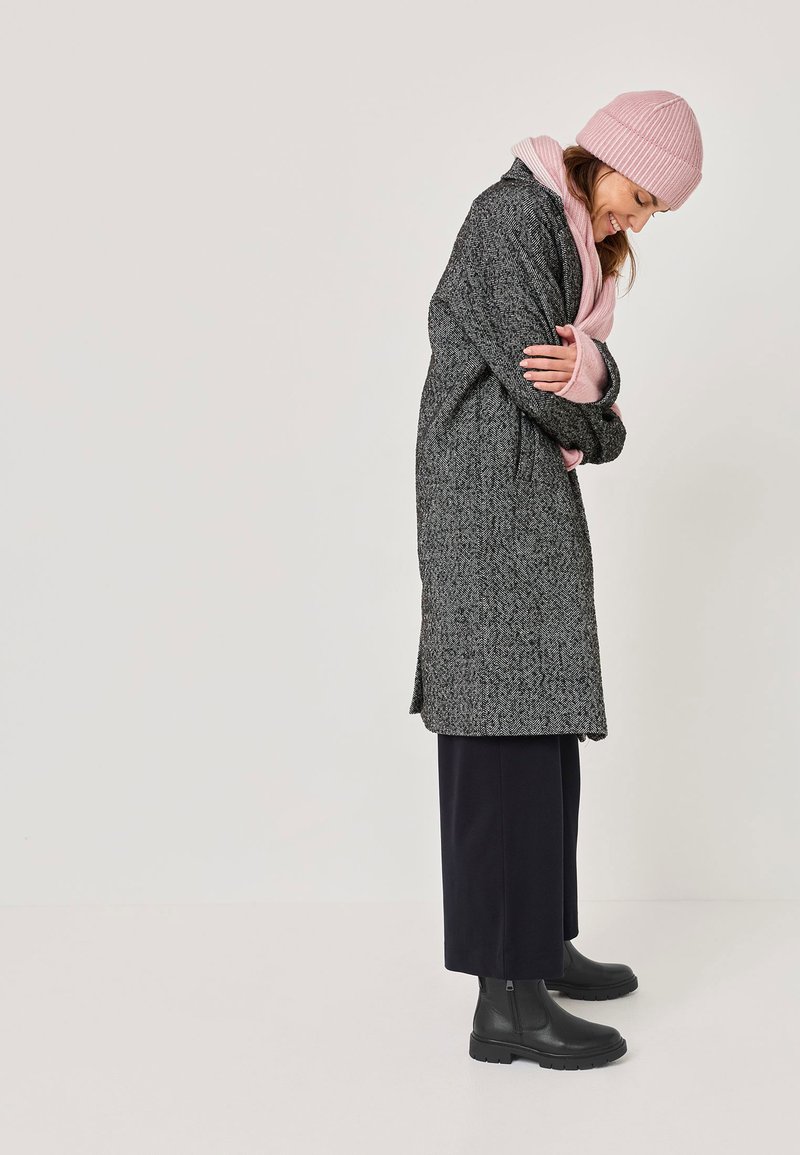 Schwarz-weiß gemusterter langer Mantel, kombiniert mit einer hellrosa gerippten Mütze und einem Schal, gestylt mit dunklen Hosen und schwarzen Stiefeletten.