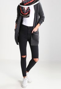 Šedý pletený cardigan s kapsami, bílý přiléhavý top, roztrhané černé skinny jeans a bílé tenisky. Šála má geometrické vzory v červené, černé a bílé.