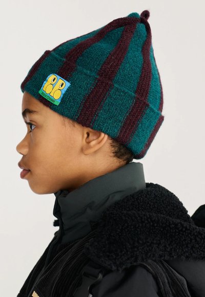 Enfant portant un bonnet tricoté rayé bleu sarcelle et violet foncé avec un patch coloré, habillé d'une veste noire avec un col en polaire, tourné vers la gauche.