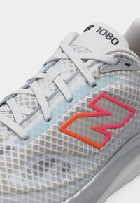 Κοντινό πλάνο άσπρου αθλητικού παπουτσιού με σχέδιο από δίχτυ, λευκά κορδόνια, λογότυπο "N" σε κόκκινο και ροζ χρώμα, και τον αριθμό "1080" τυπωμένο στη γλώσσα του παπουτσιού.