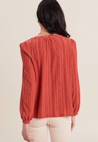Blouse rouge texturée avec des rayures verticales, des manches ballon et un dos froncé. Présente des détails brodés et une coupe décontractée.
