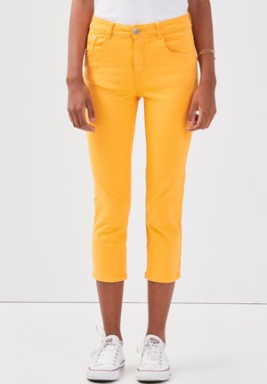 Personne portant un pantalon court jaune vif, des baskets blanches et un haut blanc, debout devant un fond blanc uni.