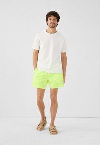 Camiseta blanca, pantalones cortos de baño amarillos neón, diseño simple, ajuste casual, tela suave, complementados con sandalias marrones con tiras texturizadas.
