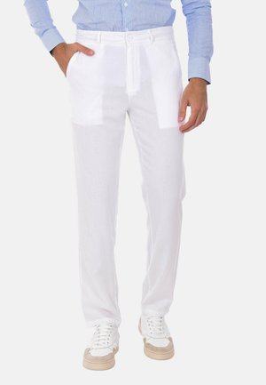 Uomo che indossa pantaloni bianchi con coulisse, camicia blu a maniche lunghe e scarpe da ginnastica bianche, in piedi con una mano in tasca.