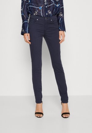 Nainen pukeutuneena tummansinisiin kapealestisiin housuihin, kuvioituun siniseen paitaan, jossa on ankkureita ja köysiä, sekä mustiin avokärkisiin korkokenkiin, seisomassa yksinkertaista taustaa vasten.