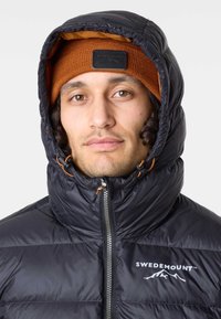 Svart vadderad jacka med hög krage, orange foder och logotyp. Åtföljs av en ribbad brun mössa. Har dragkedja och justerbara snoddar.