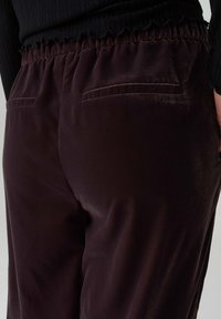Burgundy pants with a soft texture, featuring an elastic waistband and two back pockets, captured from a side angle.