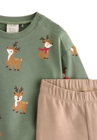 Zielony długi rękaw bluzka z nadrukiem renifera, jasnobeżowe spodnie. Bluzka ma prążkowany kołnierz i napy; spodnie z elastycznym paskiem.