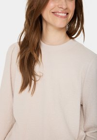 Sweatshirt de culoare roz deschis, cu un model floral texturat, guler rotund cu nervuri și croială relaxată. Materialul pare moale și plăcut la atingere.