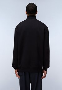 Hombre vestido con un sudadera negra de cuello alto y pantalones oscuros, de pie de espaldas contra un fondo claro y liso.