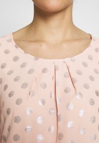 Blusa con escote redondo, hecha de una tela de color rosa claro. Presenta puntos metálicos plateados y un pliegue fruncido en el centro delantero.