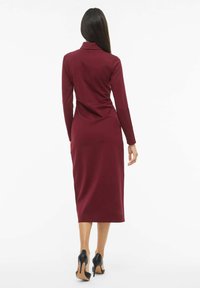 Vyšninių spalvų figūrą prigludusi suknelė ilgomis rankovėmis ir apykakle, siekianti kelių ilgio. Dėvima su juodais aukštakulniais batais, švelnios audinio tekstūros.