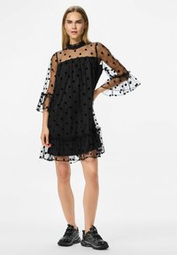 Crna haljina s točkicama od prozirnog materijala, s raširenim rukavima i naboranim rubom. Zajedno s crnim atletskim cipelama, model stoji protiv bijele pozadine.