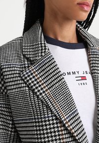 Svartvit hundtandsmönstrad kavaj med hakslag, kombinerad med en grå T-shirt med logotyp och blå halsringning.