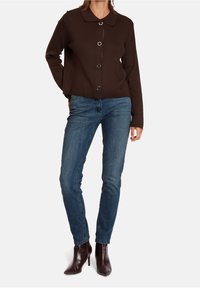 Kvinna som bär en mörkbrun knäpp cardigan, blå figursydda jeans och mörkbruna spetsiga klackboots som slutar vid vristen, stående med en hand bakom ryggen.