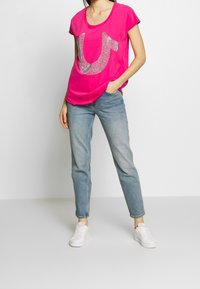 Mujer vestida con una camiseta de manga corta rosa brillante con un logo plateado, jeans azul claros y zapatillas blancas, posando con una mano en el bolsillo.