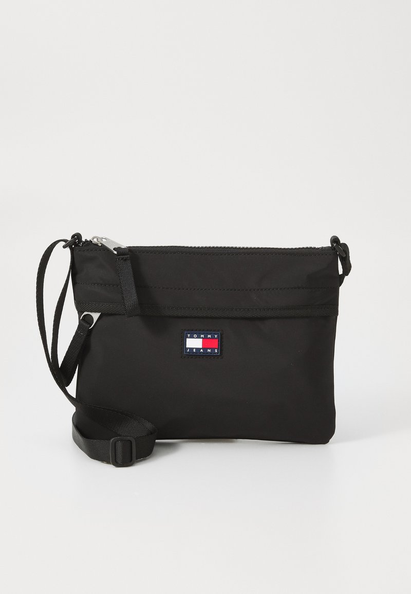 Bolsa de ombro preta em nylon com fecho de zíper, alça ajustável e um emblema retangular com apontamentos em vermelho, branco e azul.