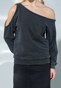 Sweatshirt gris foncé avec un design épaules dénudées, bords effilochés et poignets côtelés. Le matériau semble doux avec une coupe décontractée.