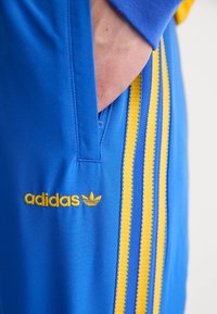 Pantalones cortos deportivos azules con la marca adidas en amarillo. Rayas laterales en amarillo y azul, textura suave y detalle de bolsillo con cremallera.