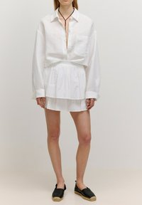 Witte linnen oversized shirt met lange mouwen, gecombineerd met een gelaagde witte rok met verticale strepen. Zwarte espadrilleslippers.