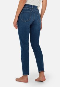 Jeans de mezclilla, de talle alto, azul oscuro, ajustados, con dos bolsillos traseros y un detalle de costura minimalista. Modelo descalzo.