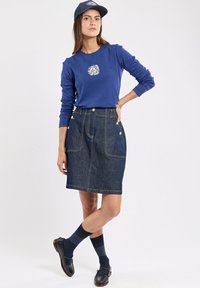 Niebieska koszulka z długim rękawem z okrągłą grafiką, w połączeniu z dżinsową spódnicą do kolan z złotymi guzikami i dużymi kieszeniami. Ciemne skarpety i buty.