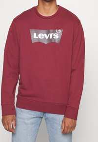 Hombre vestido con un sudadera color burdeos de Levi's con logo blanco y jeans de mezclilla azul claro, de pie contra un fondo liso.