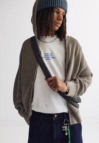 Beige Strickjacke mit Kapuze, über einem weißen T-Shirt mit Grafik, dunklen Jeans und einer Mütze getragen. Auffällige Ketten- und Schlüsselanhängerdetails.
