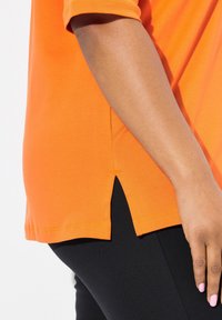 Oranje katoenen t-shirt met korte mouwen en een split aan elke kant. De stof is glad met een effen kleur, gecombineerd met zwarte leggings.