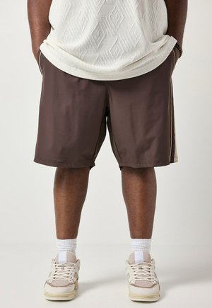 Person iført brune shorts, hvid mønstret top, hvide sokker og beige sneakers, stående med hænderne i lommerne.