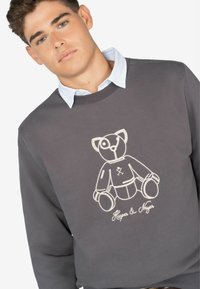 Grijze sweatshirt met een wit geborduurd teddybeerontwerp en de tekst "Harper & Nyer" aan de voorkant, met een ronde hals en textiel met structuur.