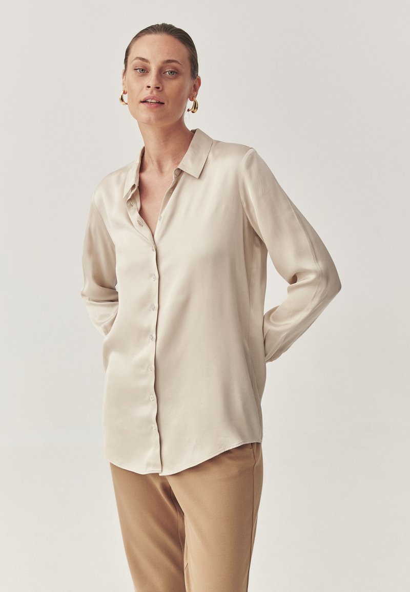 Langærmet, lys beige bluse lavet af satin, med klassisk krave og knaplukning, parret med brune bukser.