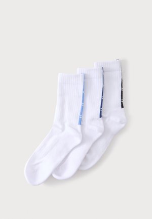 Trois paires de chaussettes blanches montantes avec des bandes colorées le long des côtés en bleu clair, bleu foncé et noir, posées en chevauchement sur un fond clair.