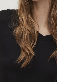 Svart topp med v-ringning, tillverkad av mjukt tyg med en slät textur; l ångt, vågigt brunt hår faller ner och delvis täcker plagget.