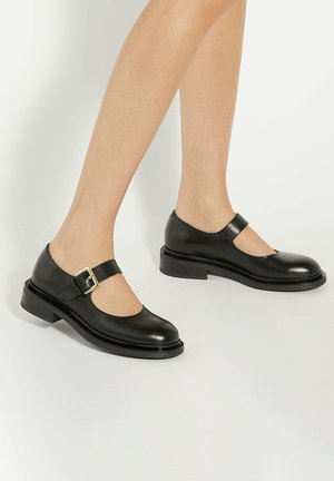 Zwarte leren Mary Jane schoenen met lage blokhakken en gouden gesps, gedragen op blote voeten tegen een effen lichte achtergrond.