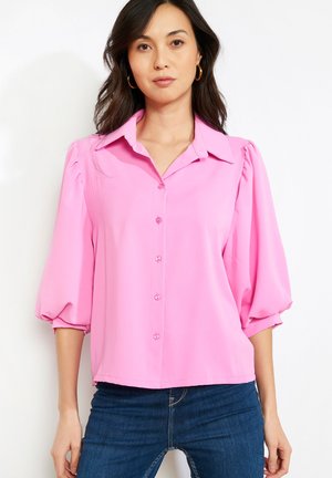 Žena nosiaca jasnoružovú blúzku na gombíky s nafúknutými tromi štvrtinovými rukávmi a modré rifle, stojaca pred bielym pozadím.