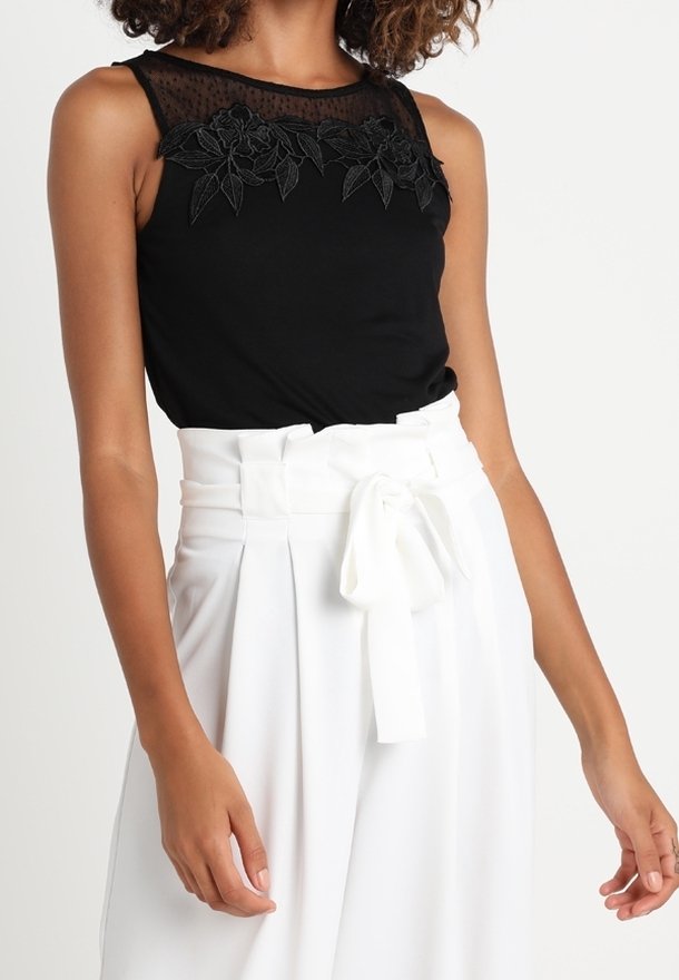 Haut noir sans manches avec des détails en dentelle florale au niveau du col, associé à un pantalon blanc taille haute avec une ceinture en tissu nouée.