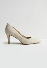 Punta di stivale beige testurizzata da donna con tacco a stiletto di media altezza, esposta su una superficie bianca contro uno sfondo chiaro e semplice.