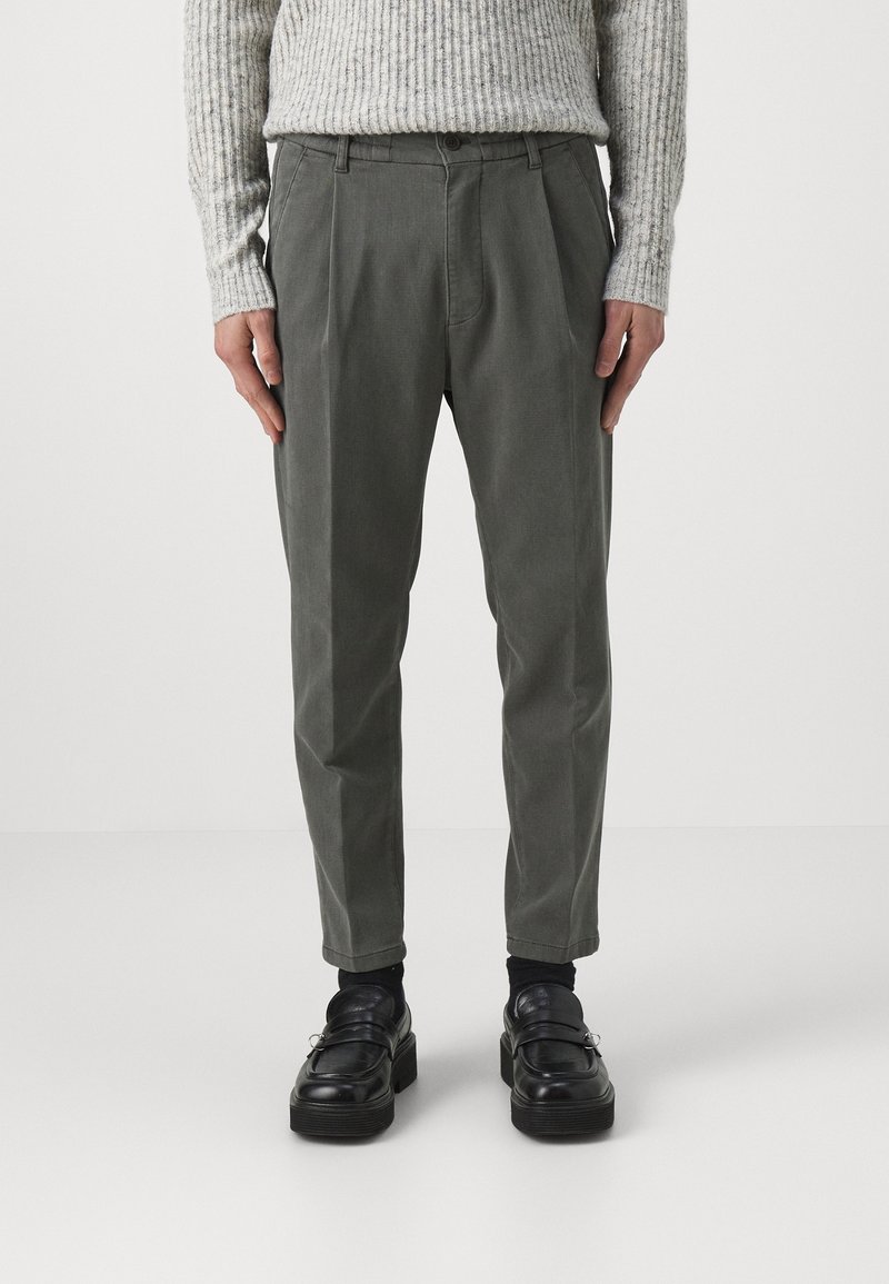 Homme portant un pantalon gris plissé, des chaussettes noires, des mocassins en cuir noir et un pull côtelé gris clair, debout les mains le long du corps.